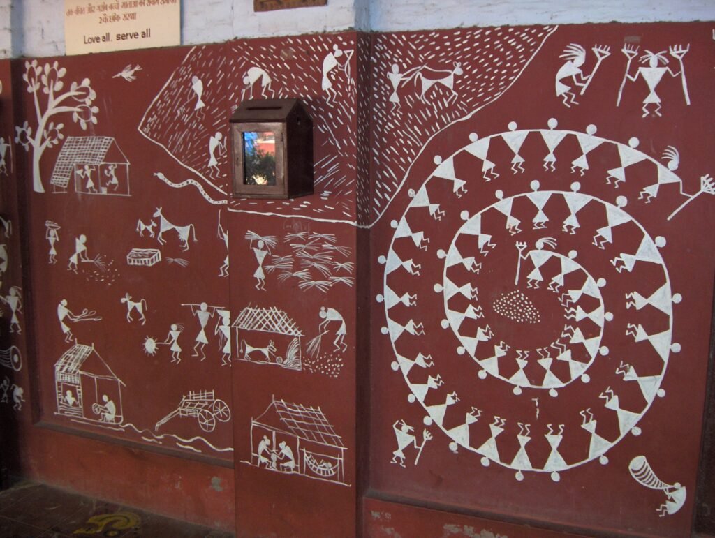 Warli tribal painting: white stick figures of villagers, animals and trees on an earth-red ground